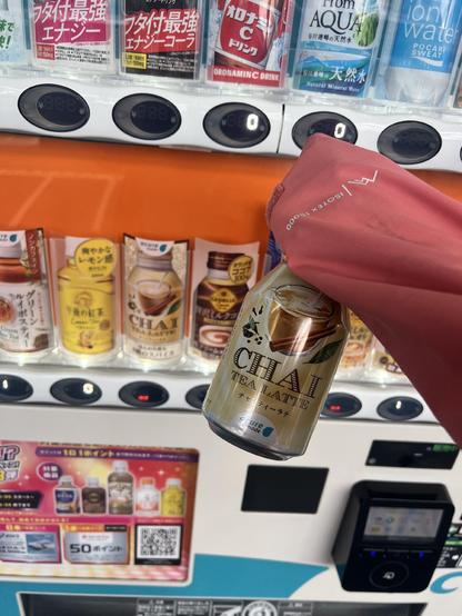 A metal bottle of chai tea held (with a hand covered with a coat sleeve) in front of a Japanese vending machine where some drinks how have a red “hot” label.