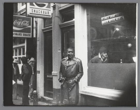 The image is a black and white photograph depicting an urban street scene. In the foreground, there's a man standing in front of what appears to be a snack bar or cafe with large glass windows reflecting the surrounding environment. He is wearing a leather coat, has his hands casually placed on his hips, and seems engaged in conversation as suggested by his open mouth and expressive facial features.

The establishment he stands before bears signs for "Coca-Cola" and possibly another beverage brand like "Dewars Snack," indicating it could be a snack bar or cafe. The architecture suggests an older style of building with large glass windows, typical of urban commercial buildings in the mid-20th century.

In the background through the window reflection, there are glimpses of two other individuals; one appears to be inside and seated at a table, while another is outside on what seems like a city street. The overall atmosphere gives off a candid or snapshot-like quality, capturing a slice of life moment in an urban setting.

This photograph carries historical value as it's part of the collection by Ed van der Elsken, known for his work documenting various aspects of Dutch culture and daily life during different periods. This specific image likely falls within one such documentation period between 1975-1990 at Zeedijk in Amsterdam.